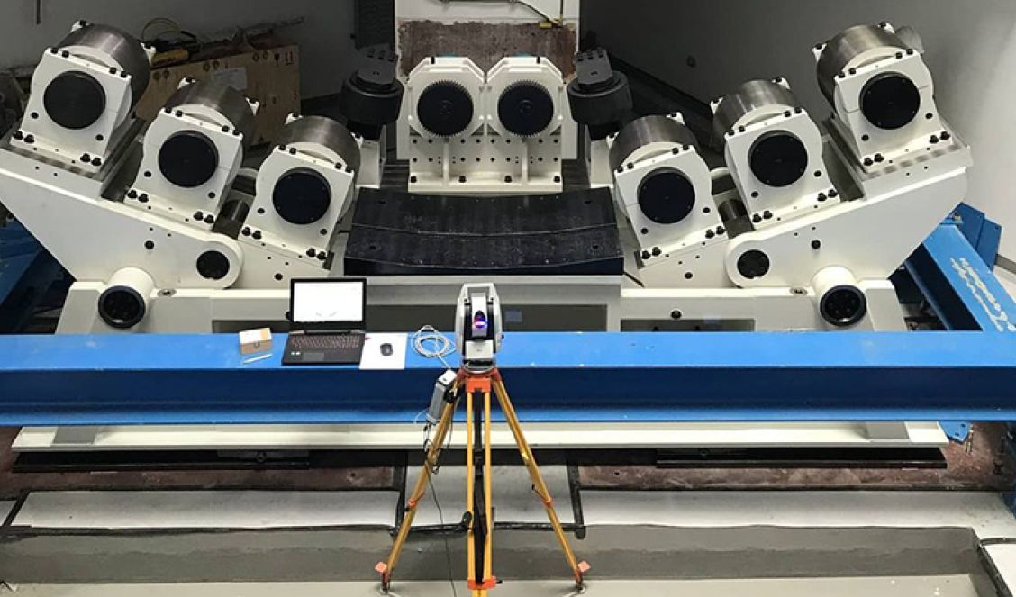 A large industrial machine with multiple rotating arms is positioned on a blue metal frame. In front, a tripod-mounted measuring device, such as the Leica AT960 for laser tracker alignment, alongside a laptop and keyboard are set up for calibration or testing.