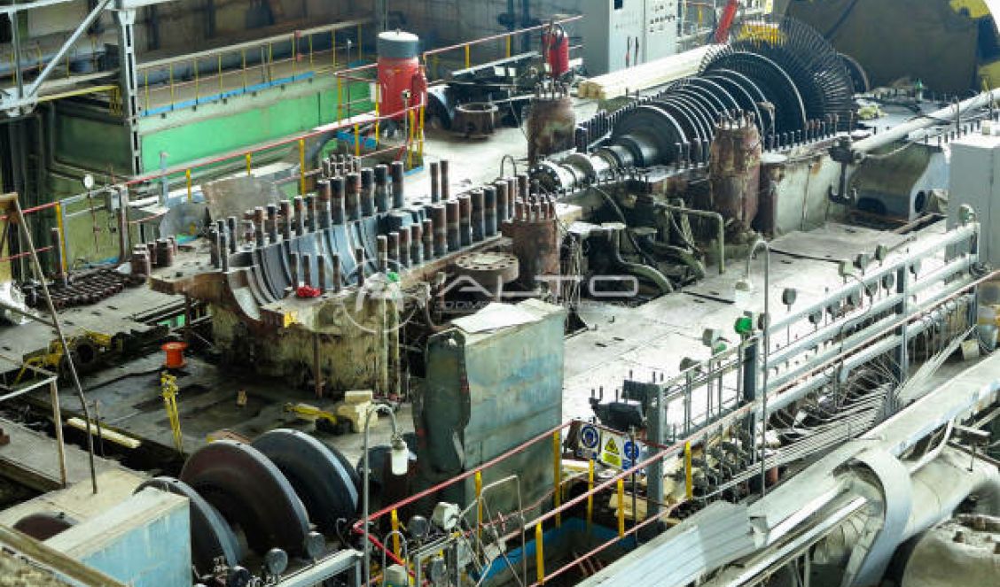 A massive disassembled turbine rotor resting horizontally on supports within a large industrial workshop hall.